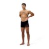 גבר מודרני לובש בגד ים שחייה של Speedo, עומד על רקע לבן.